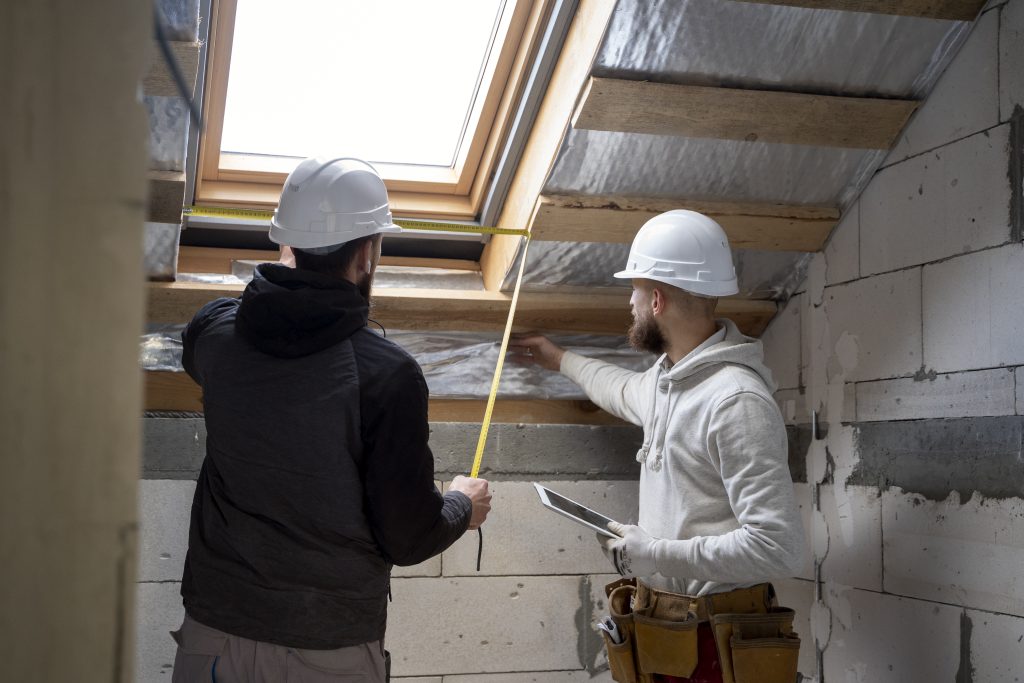 Two construction workers wearing white hard hats measuring and inspecting a loft space under renovation. One holds a measuring tape while the other uses a tablet, discussing insulation and structural elements