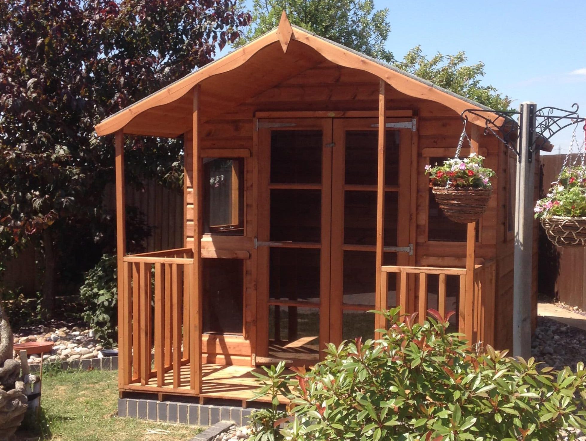 Wooden garden shed with porch, surrounded by plants and hanging baskets, under a clear blue sky.