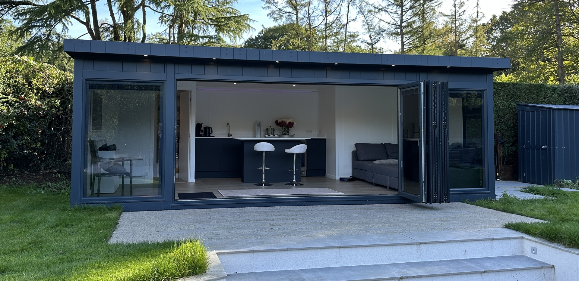 Modern garden office with open bi-fold doors, featuring a stylish interior, bar stools, and surrounded by lush greenery