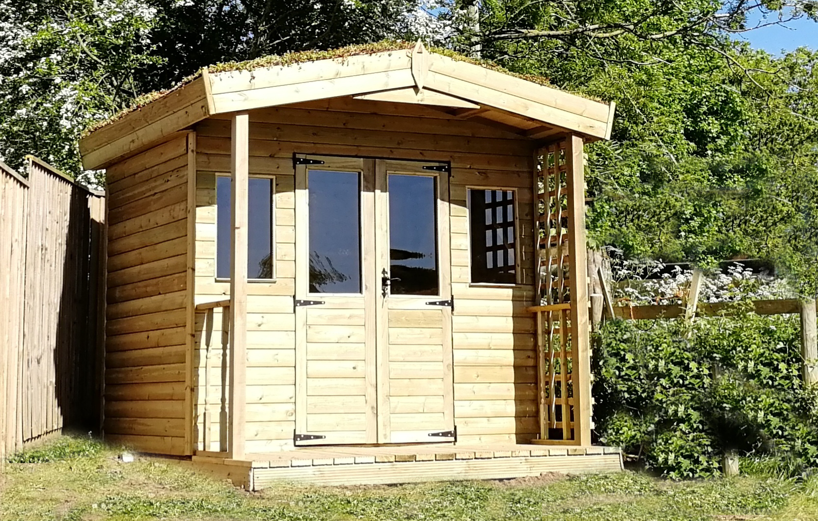 Wooden garden shed with glass doors, surrounded by greenery, under bright sunlight